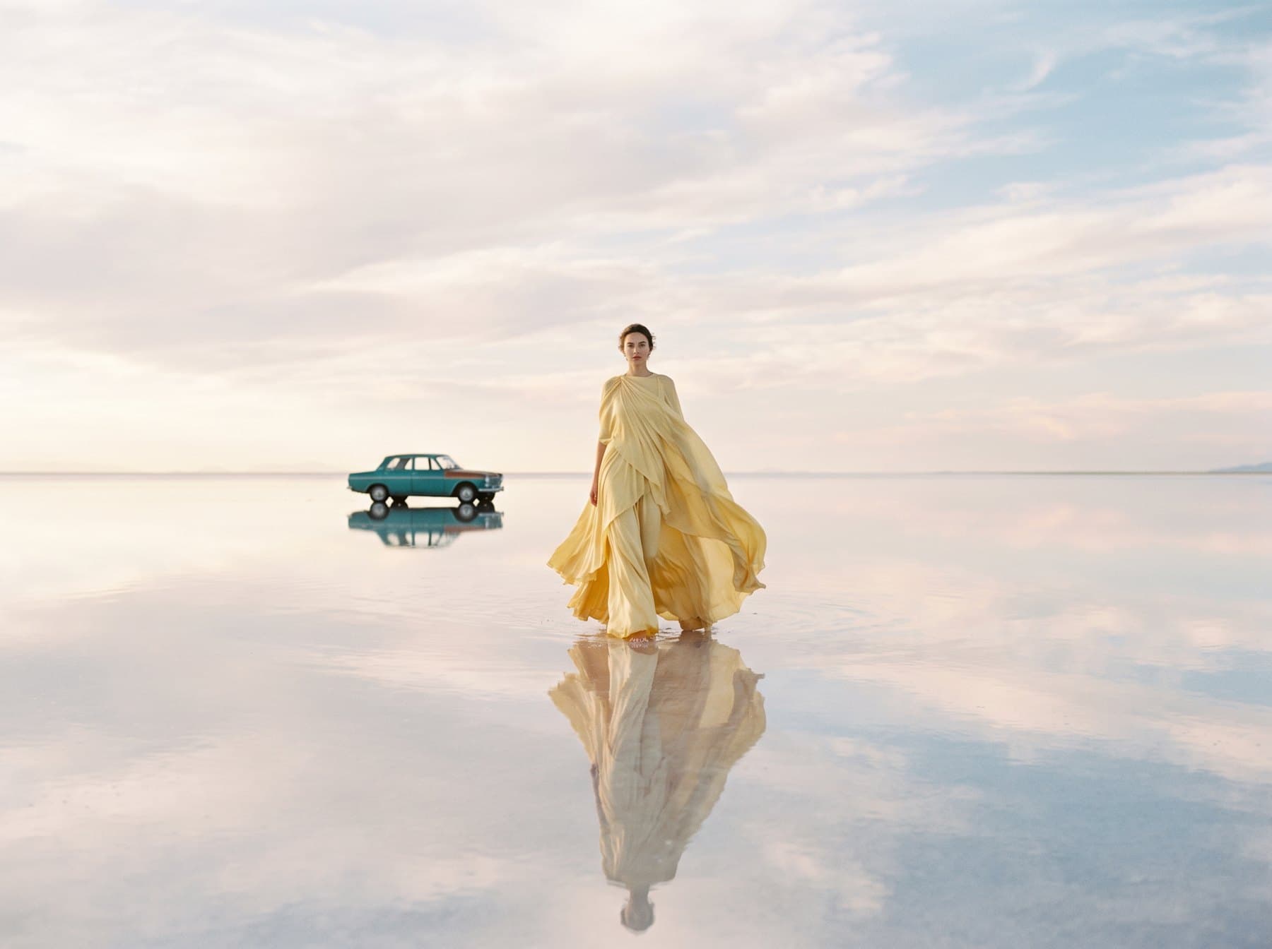 A woman in a flowing yellow gown on a mirror-like salt flat, a vintage car behind her